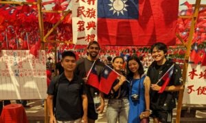 Students stand in front of a red flag with a blue square and a yellow sun symbol in the middle. A student holds the same flag in their hand.