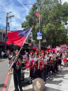 A group of military individuals sit down in the middle of the street and one man waves a red flag with a blue square and a white sun symbol.