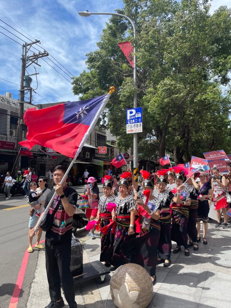 A group of military individuals sit down in the middle of the street and one man waves a red flag with a blue square and a white sun symbol.