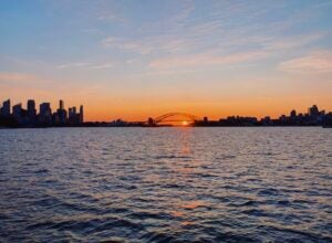 The skyline of Sydney, Australia at sunset illuminates hues of orange, yellow, and red against a partly-cloudy sky.