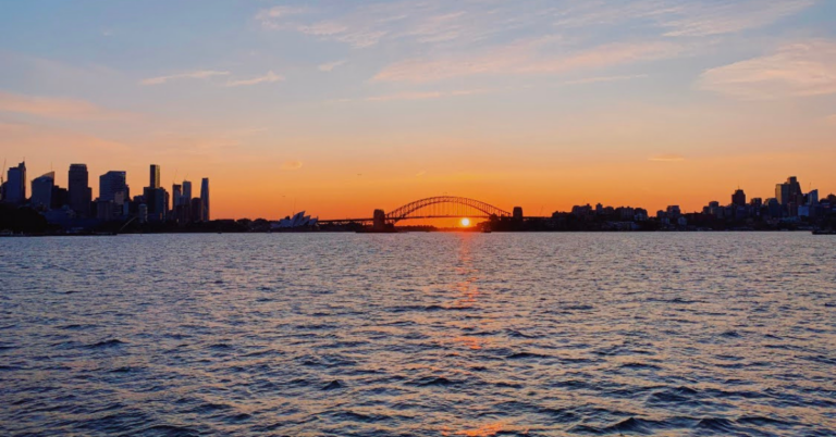 The skyline of Sydney, Australia at sunset illuminates hues of orange, yellow, and red against a partly-cloudy sky.