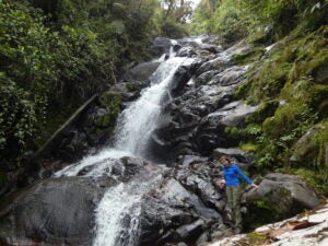 Student in blue raincoat capturing a photo at the base of a waterfall.