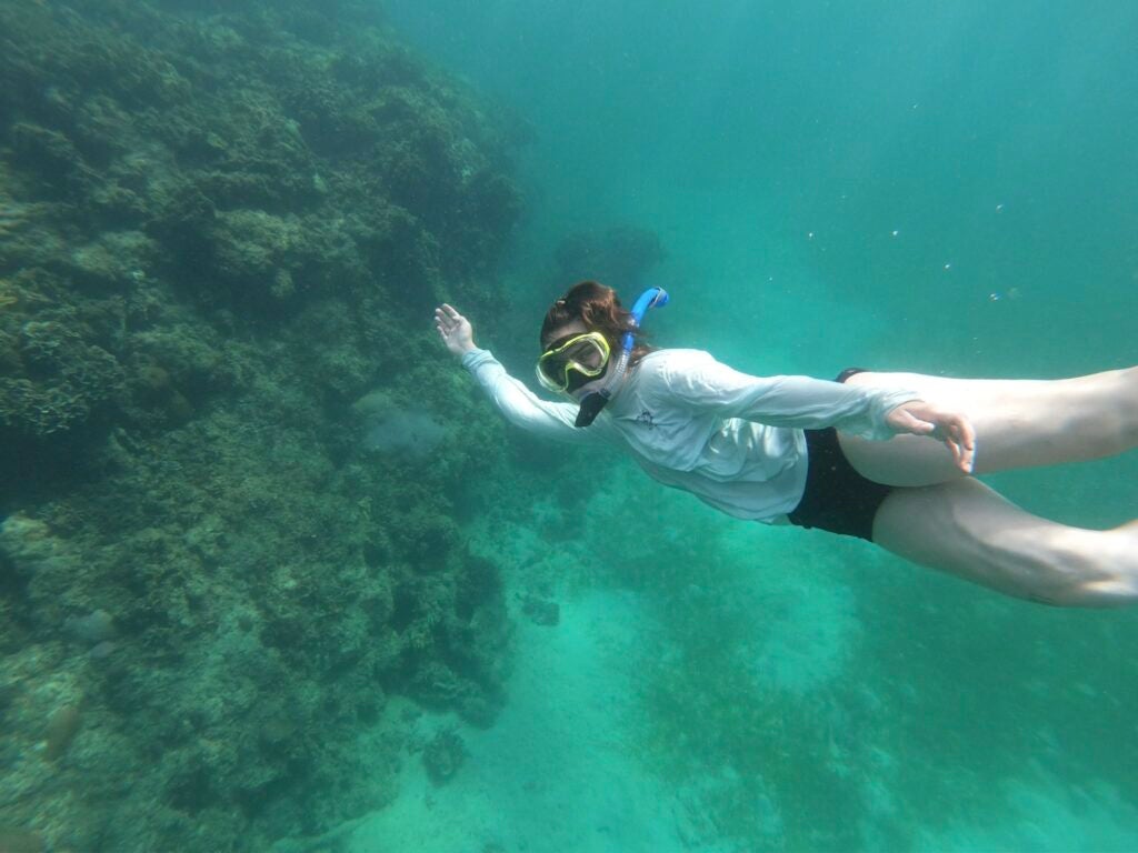 A young lady is snorkeling and smiling at the camera. She wears a white. long sleeve top and dark bottoms while underwater with light snorkeling gear.