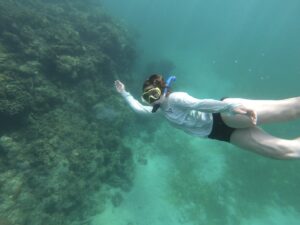 A young lady is snorkeling and smiling at the camera. She wears a white. long sleeve top and dark bottoms while underwater with light snorkeling gear.