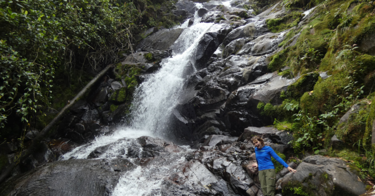 Student in blue raincoat capturing a photo at the base of a waterfall.