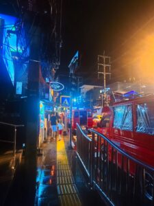 The busy night streets of Bangkok show people walking around a brightly lit city center with neon building signs.