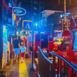The busy night streets of Bangkok show people walking around a brightly lit city center with neon building signs.