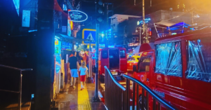 The busy night streets of Bangkok show people walking around a brightly lit city center with neon building signs.
