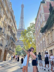 Two students stand back to back with one arm up in the air drawing the attention to the Eiffel Tower behind them while on a busy street in Paris.