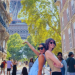 Two students stand back to back with one arm up in the air drawing the attention to the Eiffel Tower behind them while on a busy street in Paris.