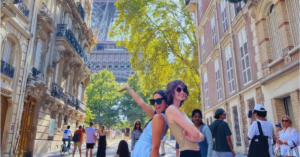 Two students stand back to back with one arm up in the air drawing the attention to the Eiffel Tower behind them while on a busy street in Paris.