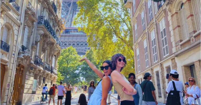 Two students stand back to back with one arm up in the air drawing the attention to the Eiffel Tower behind them while on a busy street in Paris.