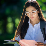 Female student in white glasses and a white top studies a booklet while walking outside on a sunny day.