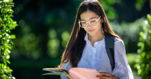 Female student in white glasses and a white top studies a booklet while walking outside on a sunny day.