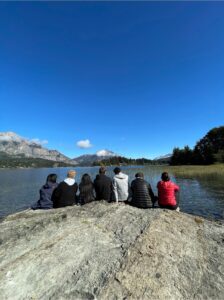A group of students sit on a rock in front of a large lake and a mountain with snow in the distance. 