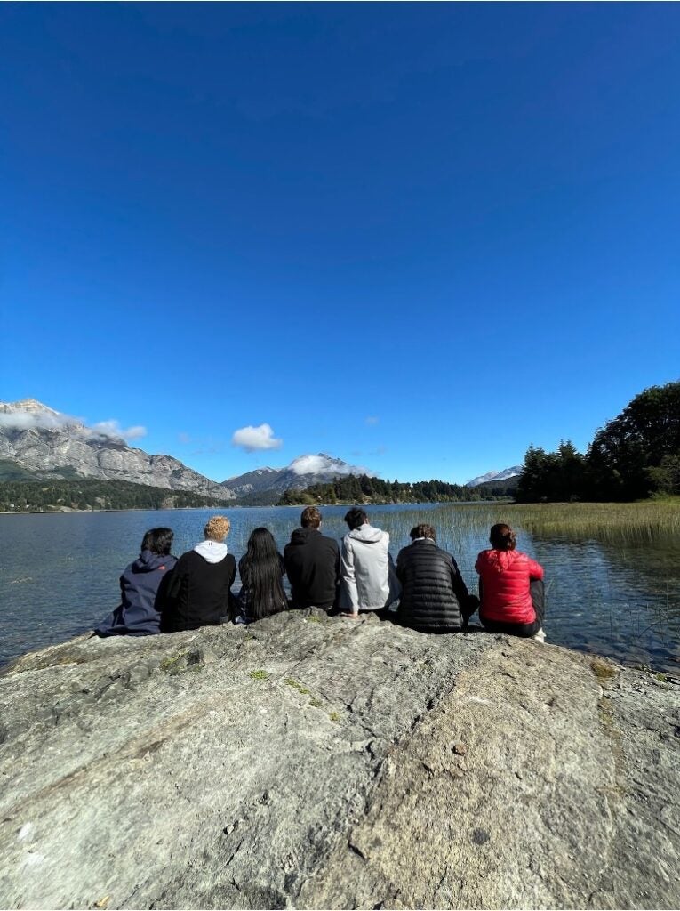 A group of students sit on a rock in front of a large lake and a mountain with snow in the distance.