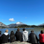A group of students sit on a rock in front of a large lake and a mountain with snow in the distance.