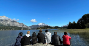 A group of students sit on a rock in front of a large lake and a mountain with snow in the distance.
