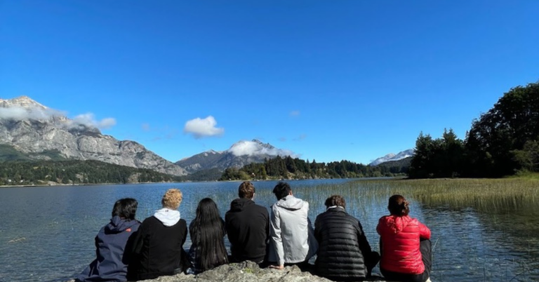 A group of students sit on a rock in front of a large lake and a mountain with snow in the distance.