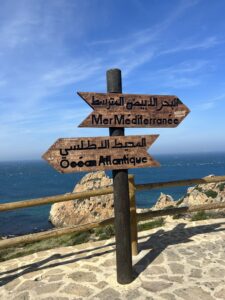 Wooden signs in Arabic facing two different ways on a highway overlooking a clear, blue sky and an ocean.