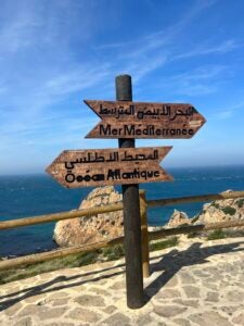 Wooden signs in Arabic facing two different ways on a highway overlooking a clear, blue sky and an ocean.