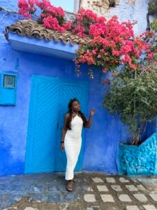 A student in a white dress stands and smiles in front of a house in bright blue. There are pink flowers decorating the house.