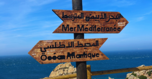 Wooden signs in Arabic facing two different ways on a highway overlooking a clear, blue sky and an ocean.