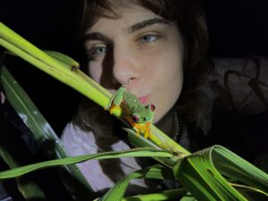 A student poses for a photo behind a frog on a long leaf outdoors at nnight.