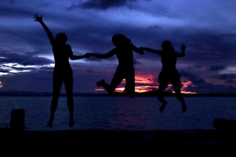 Three shadows jump up while holding hands and on the beach as the sun sets.