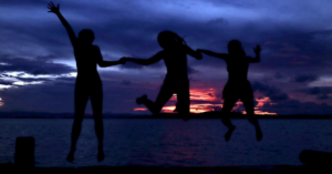 Three shadows jump up while holding hands and on the beach as the sun sets.