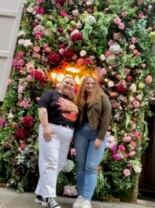 Two students smile for the camera behind a backdrop of fresh flowers and greenery. The students are wearing casual dress with jeans, t-shirts and jackets.