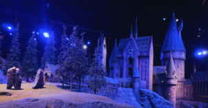 A caled version of the exterior of the Hogwarts castle at Harry Potter Studios in London is lit in hues of dark blue and black. The building is snowy and looks like a magical winter wonderland.