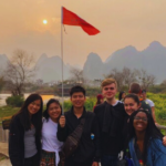 A group of seven students pose for a photo on a hazy orange day with a mountain range behind them and a red flag flying high.
