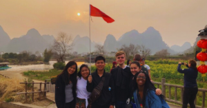 A group of seven students pose for a photo on a hazy orange day with a mountain range behind them and a red flag flying high.