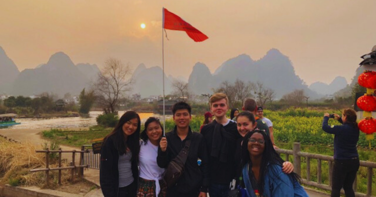 A group of seven students pose for a photo on a hazy orange day with a mountain range behind them and a red flag flying high.
