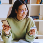 A girl in a green sweater is looking at her credit card with a phone in her hand. She is surrounded by books and a bookshelf.