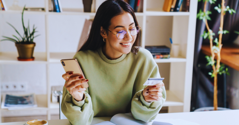 A girl in a green sweater is looking at her credit card with a phone in her hand. She is surrounded by books and a bookshelf.