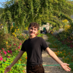 A student wearing a black t-shirt and black denim smiles at the camera with both hands held out wide at their side. They are in a garden of lush greenery and colorful flowers.