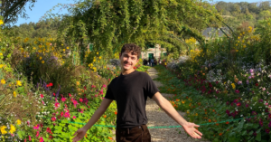 A student wearing a black t-shirt and black denim smiles at the camera with both hands held out wide at their side. They are in a garden of lush greenery and colorful flowers.