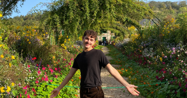 A student wearing a black t-shirt and black denim smiles at the camera with both hands held out wide at their side. They are in a garden of lush greenery and colorful flowers.