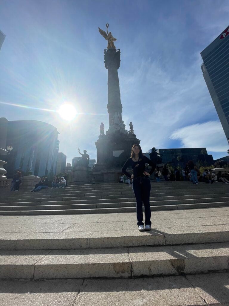 A young lady stands in front of a large angel sculpture in a plaza.