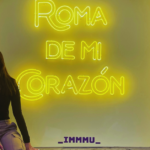 A young woman sitting on a ledge, facing the camera, with an illuminated window or sign reading 'roma de mi corazon' behind her.