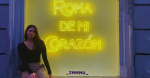A young woman sitting on a ledge, facing the camera, with an illuminated window or sign reading 'roma de mi corazon' behind her.