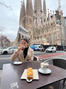 A student in a black trench coat looks at the camera while sipping a small cup of hot coffee in the city center with large, historic buildings behind her.