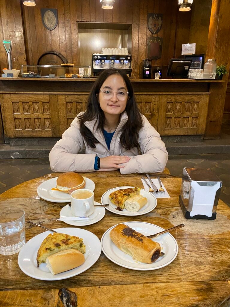 Student with glasses wearing a light pink sweater smiles at the camera while seated at a dinner table with many food options in front of her