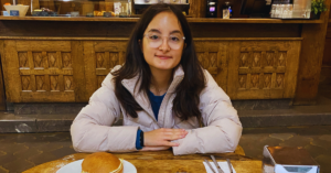 Student with glasses wearing a light pink sweater smiles at the camera while seated at a dinner table