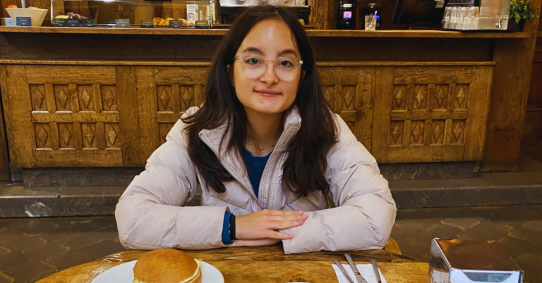 Student with glasses wearing a light pink sweater smiles at the camera while seated at a dinner table