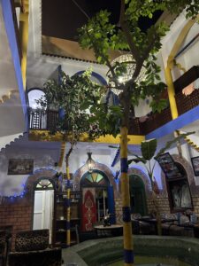 The inside of a hostel in Morocco has two stories, an outdoor plaza, a wrap around patio on the first and second floors and a tree in the middle.