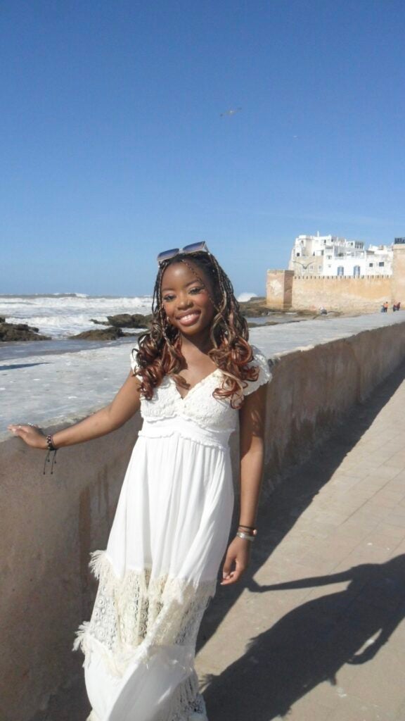 A student in a white dress smiles while putting one hand on a wall in an old town or city.