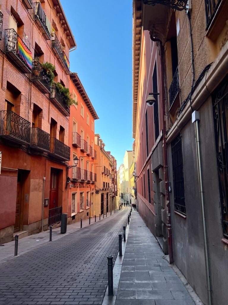 The bright orange and pink buildings on either side of a paved street house small balconies made of iron rods on each level.